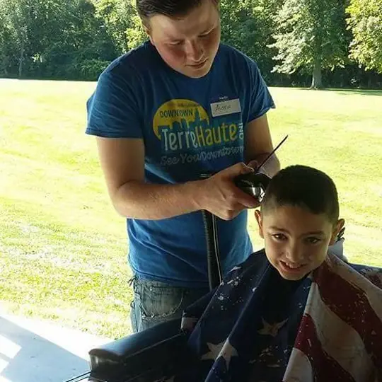 A barber giving a boy a haircut outdoors on a sunny lawn, the boy in a stars-and-stripes cape